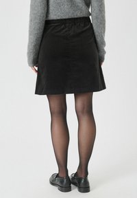 Zwarte corduroy rok met een uitlopende silhoutte en elastische tailleband, gecombineerd met doorschijnende panty’s en zwarte loafers.