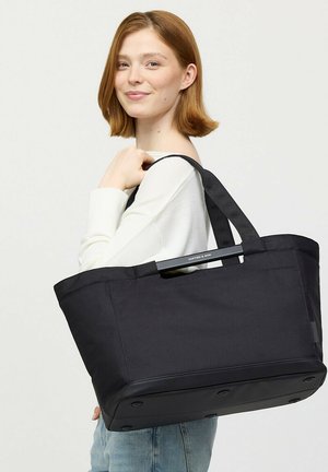 Junge Frau mit kurzen roten Haaren trägt eine große schwarze Tragetasche über der Schulter vor schlichtem Hintergrund.