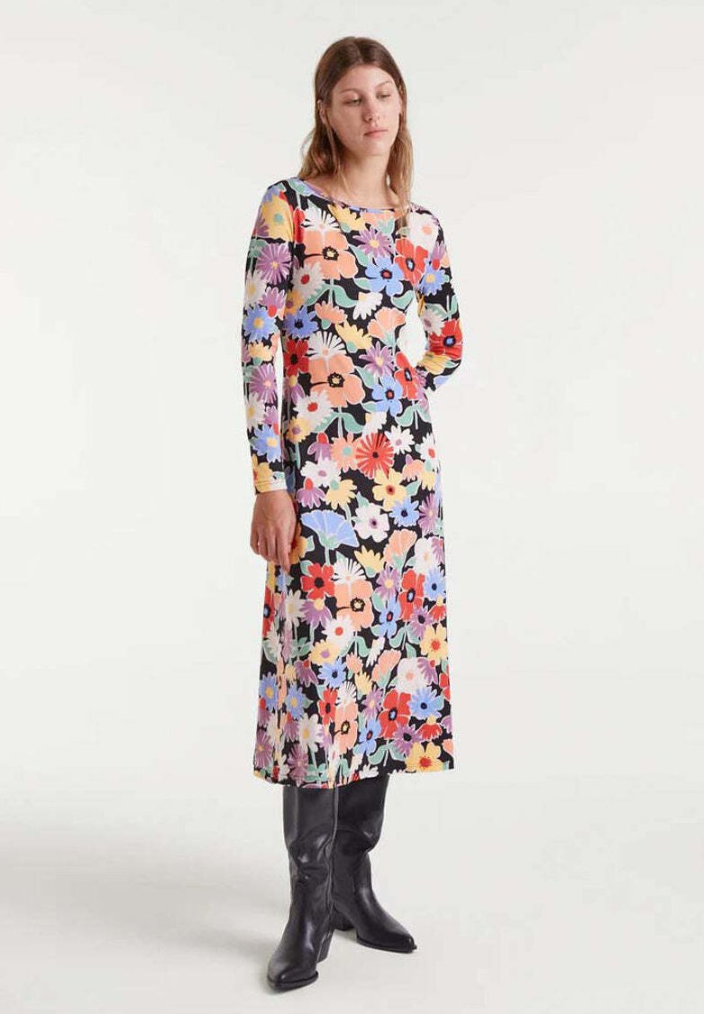 Mujer vistiendo un vestido floral de manga larga y largo hasta los tobillos con fondo negro y botas negras hasta la rodilla, frente a un fondo blanco liso.