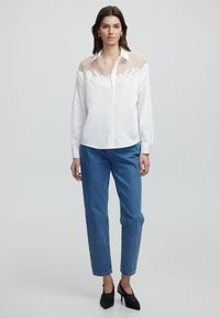 Weißes Hemd mit Knöpfen und Netzdesign an den Schultern, kombiniert mit blauen Jeans und schwarzen hohe Absätzen. Glatte Stoffstruktur.