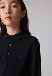 Must must ümmargune krae, kolme nupuga ja väikese tikandiga logoga vasakul rinnal, valmistatud pehmest kangast must polo särk.