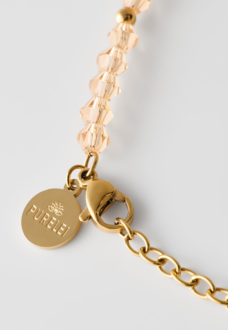 Goldfarbene Kettenarmband mit pfirsichfarbenen Kristallperlen und einem runden Anhänger, der mit "PURELEI" graviert ist, gesichert mit einem Karabinerverschluss.