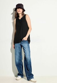 Top nero senza maniche con una finitura texturizzata, abbinato a jeans a gamba larga blu e sneaker bianche. La persona indossa un cappello a secchiello nero.