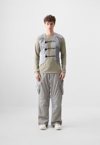 Grå vadderad väst med svarta knappar, bärs över en långärmad olivfärgad skjorta, tillsammans med lösa grå cargobyxor och benvita sneakers.