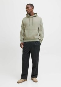 Felpa khaki con tasca frontale e cordino regolabile, abbinata a pantaloni plissettati scuri e calzature beige. Tessuto liscio.