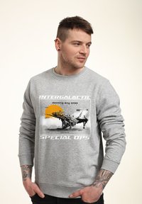 Szary sweatshirt z graficznym nadrukiem statku kosmicznego, z żółtymi i czarnymi akcentami. Żebrowane mankiety i pas. Luźny krój.