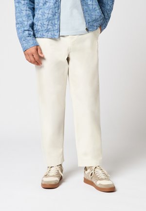 Homme portant un pantalon droit écru, des baskets beige et blanches, une chemise bleu clair et une veste bleue à motifs, debout les mains dans les poches.