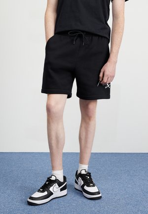 Personne portant un short noir à cordon, des baskets noir et blanc, et des chaussettes blanches, debout sur un tapis bleu sur fond uni.