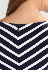 Marinblå och vitrandig topp med ett chevronmönster och en detalj i nacklinningen bak som är fäst med en liten mässingsknapp. Mjukt, texturerat tyg.