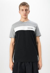 Ung mand iført en farveblok T-shirt med grå ærmer, hvid bryststribe, sort krop og "RIP CURL" logo på brystet, der står foran en hvid baggrund.