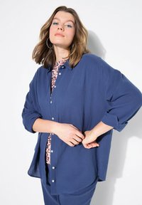 Camicia blu navy con bottoni, realizzata in tessuto testurizzato, maniche lunghe arrotolate fino ai gomiti, caratterizzata da un design a strati con uno strato sottostante con motivo.