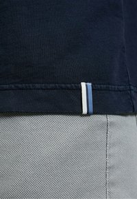 Marineblaues Baumwoll-T-Shirt mit strukturierter Oberfläche; Saum mit einem weißen und blauen gestreiften Stoffetikett. Grauer gemusterter Stoff darunter.