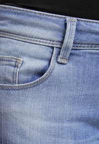 Jeans de denim azul claro con un bolsillo frontal, ligero desgaste y costuras amarillas contrastantes a lo largo de las costuras y la cinturilla.