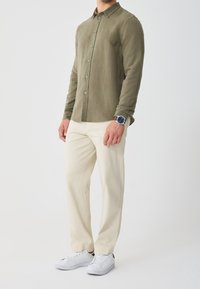 Olivově zelená košile na knoflíky s dlouhými rukávy, v kombinaci s krémovými kalhotami a bílými teniskami. Hodinky na zápěstí, ležérní střih.