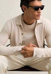 Cardigan bej deschis cu butoane argintii, purtat peste o cămașă asortată. Modelul poartă ochelari de soare negri, așezat pe un scaun.