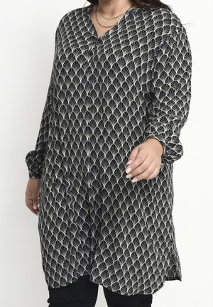 Vrouw die een lange zwarte tuniek draagt met witte waaierachtige patronen en losse lange mouwen, gecombineerd met zwarte broek en eenvoudige gouden kettingen.