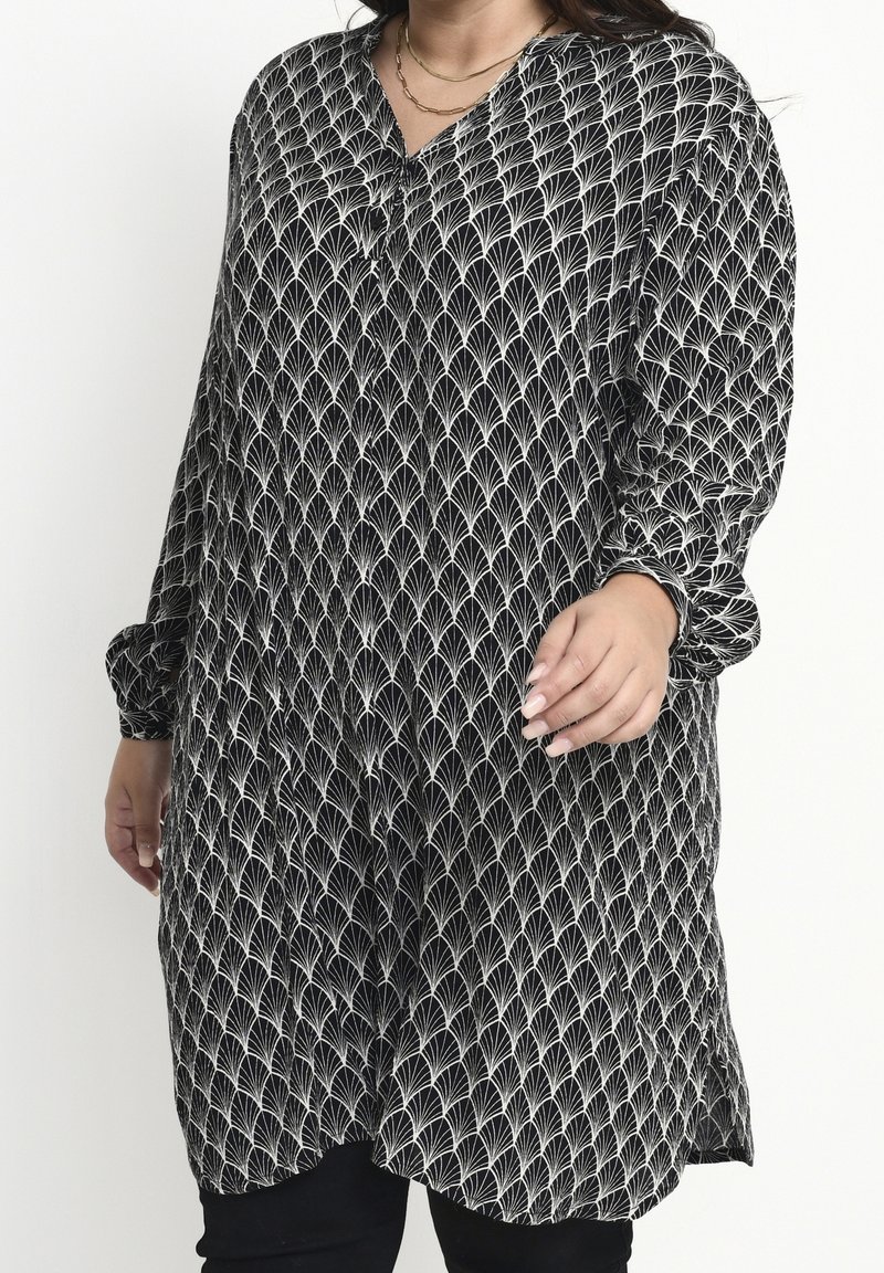 Femme portant une longue tunique noire avec des motifs blancs en forme d'éventail et des manches longues amples, associée à un pantalon noir et de simples colliers en or.