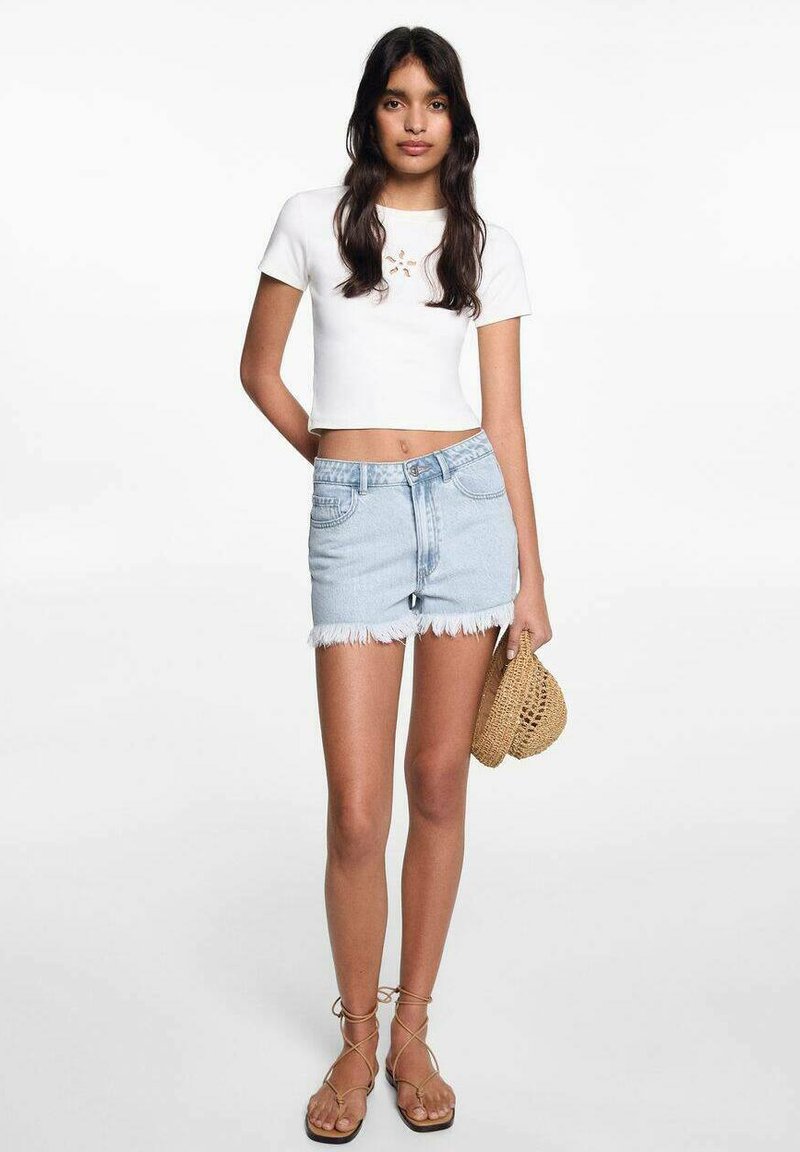 Mlada žena u bijelom kratkom topu, svijetlim traper šortsevima s rubovima, sandalama na vezanje, drži pleteni slamasti šešir ispred bijele pozadine.