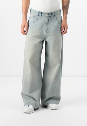 Personne portant un jean large bleu clair, des baskets blanches et un débardeur blanc, debout devant un fond uni.