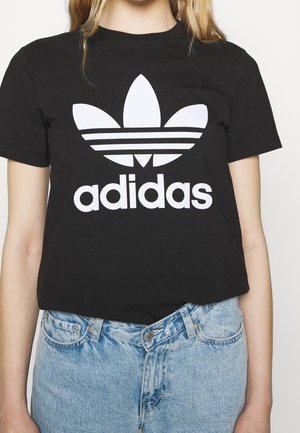Osoba ubrana w czarną koszulkę Adidas z dużym białym logo i jasnoniebieskie dżinsy, pokazana od szyi do bioder.