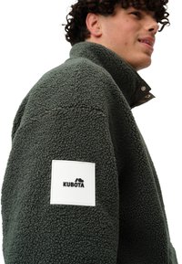 Veste en polaire vert foncé avec un col haut, un tissu texturé et une étiquette brodée blanche portant le nom "KUBOTA" sur la manche gauche.