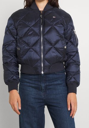 Vrouw die een marineblauwe doorgestikte bomberjas draagt met geribbelde manchetten en kraag, een rits aan de voorkant en een blauwe spijkerbroek.