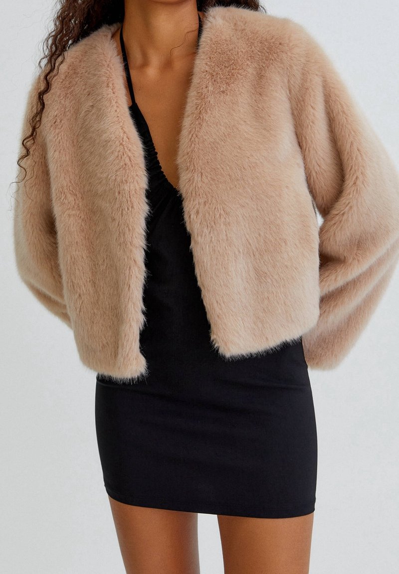 Mjuk, faux fur kort jacka i ljusbeige, med rund hals och lösa ärmar, matchad med en åtsittande svart klänning.