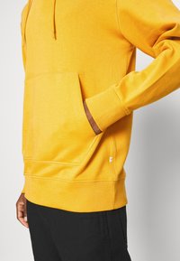 Persona vestida con una sudadera amarilla brillante con una mano dentro del bolsillo delantero, combinada con pantalones negros frente a un fondo simple.
