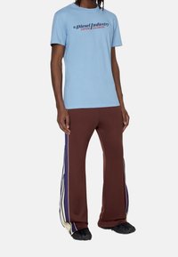 Jasnoniebieska koszulka z okrągłym dekoltem z logo "Diesel Industry" na przodzie. Brązowe spodnie dresowe z bocznymi paskami i zamkami błyskawicznymi, w połączeniu z czarnymi butami.