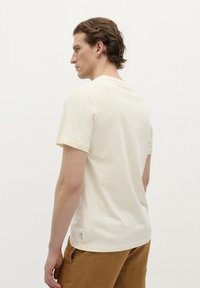Crème-kleurig katoenen T-shirt met korte mouwen, ronde hals en een klein logolabel dichtbij de zoom. Gemaakt met een paar bruine linnen broeken.