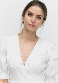 Robe blanche avec manches bouffantes, décolleté en V profond et détail de lien au niveau de la poitrine. Texture brodée et légère brillance visible.