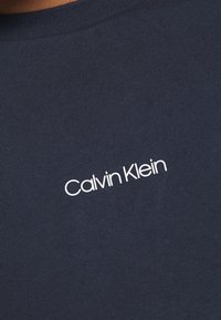 Camiseta de tela acanalada azul marino con un logo blanco de "Calvin Klein" centrado en el pecho, mangas cortas y cuello redondo.