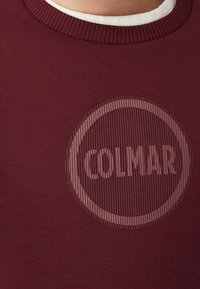 Bordeaux neulepaita, jossa on ribbauskaulus ja pyöreä logo, jossa lukee "COLMAR" keskellä.