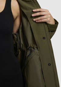 Veste imperméable verte avec un intérieur à cordon de serrage, dotée de boutons-pression et d'une texture lisse. Un débardeur noir à côtes est visible en dessous.