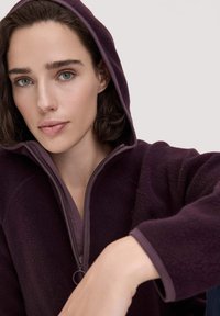 Lila Fleecejacke mit Kapuze, Reißverschluss und kontrastierenden Saumdetails. Weiche Textur, eng anliegendes Design, geeignet für die Freizeit.