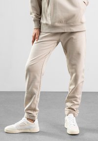 Beige joggerbukser med elastiske manchetter, sidelommer og en glat tekstur, parret med hvide sneakers med perforeret design.