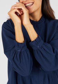 Blusa blu navy con scollatura arricciata, pattina con bottoni e maniche a palloncino voluminose. Realizzata in tessuto morbido con una sottile trama.