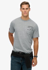 Camiseta de algodón gris con cuello redondo y mangas cortas, con un bolsillo en el pecho izquierdo y detalles de texto en negro. Llevada con un suéter oscuro atado a la cintura.