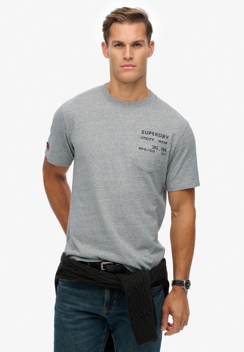 Camiseta de algodón gris con cuello redondo y mangas cortas, con un bolsillo en el pecho izquierdo y detalles de texto en negro. Llevada con un suéter oscuro atado a la cintura.