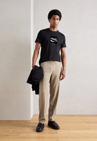 Black cotton t-shirt with Coca-Cola graphic, beige pleated trousers, and black slip-on shoes. Model holding a black jacket. Wood floor.
