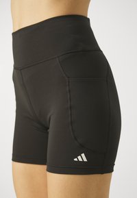 Schwarze Sportshorts aus elastischem Material, mit hohem Taillenbund, Seitentasche und weißen Logodetails am unteren Bein.