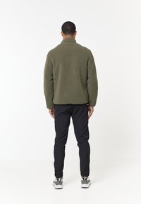 Fleecejakke i olivengrønn, med høy krage, teksturert overflate og avslappet passform, kombinert med svarte slim fit bukser og grå joggesko.