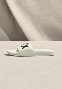 Sandália branca tipo slide com uma alça texturizada que apresenta um design de cruz preta, uma palmilha lisa e "ARMANI EXCHANGE" gravado na sola.