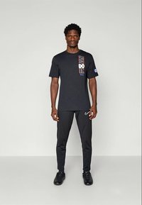 Homme debout, face à la caméra, portant un t-shirt noir Nike "Just Do It", un pantalon de jogging noir Nike avec un swoosh blanc et des baskets noires.