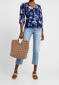 Blommönstrad marinblå blus med trekvartslånga ärmar, i kombination med ljusblå jeans och en flerfärgad vävd tote-väska med ett runt handtag.
