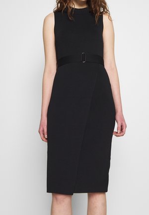 Femme portant une robe noire sans manches longueur genou avec une taille cintrée et une petite boucle de ceinture au centre.