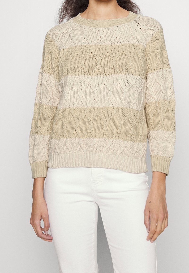Femme portant un pull en maille torsadée beige et crème à rayures avec manches longues et un pantalon blanc, vue du cou aux hanches.