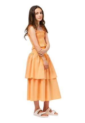 Tweedelig outfit bestaande uit een aangesloten, gesmokte oranje top en een vloeiende, gelaagde rok. Model draagt witte sandalen; de stof lijkt licht en luchtig.