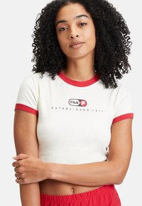 Camiseta blanca recortada con cuello y mangas acanalados rojos, que presenta un logotipo y el texto "Establecido en 1911" en negro. Textura suave.