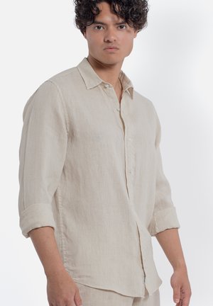 Giovane uomo con capelli ricci che indossa una camicia beige a maniche lunghe con le maniche arrotolate, in piedi davanti a uno sfondo bianco.